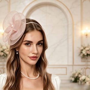 Könnte beinhalten: Ein hellrosa Fascinator-Haarreif mit einer großen, mehrlagigen, transparenten Organzablüte und einem zentralen Perlenakzent. Das Accessoire wird mit einer schlichten Perlenkette und einem einzelnen Perlenohrring kombiniert, was den sanften Look unterstreicht.