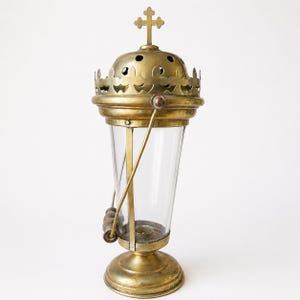 Può includere: Un incensiere in ottone con corpo in vetro trasparente, coperchio a cupola con fori circolari e un terminale a croce sulla sommità. La struttura in metallo presenta un bordo decorativo, una base circolare e un manico mobile con perline.
