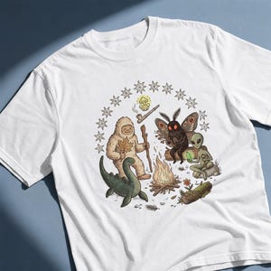Può includere: Una t-shirt bianca con un'illustrazione grafica di un Bigfoot, una creatura falena, un alieno e un mostro marino riuniti attorno a un falò. Il disegno include una pipa, un fungo e un cerchio di stelle sopra le figure.