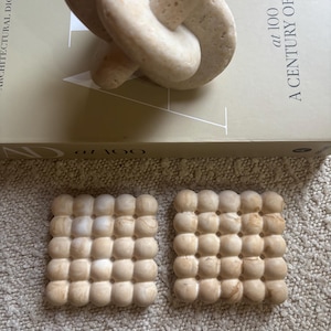 Può includere: Due sottobicchieri quadrati con una griglia di protuberanze arrotondate in rilievo con finitura in pietra marmorizzata beige, posizionati davanti a un libro intitolato Architectural Digest at 100 A Century of Style con una scultura a nodo.
