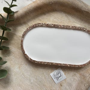 May include: An oval-shaped tray with a smooth white center and a textured rose gold glitter border. A small square white label with the text Bubble & Bond is positioned near the tray. A green eucalyptus stem is placed to the left of the tray.