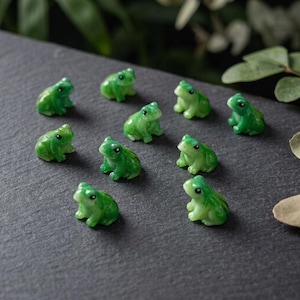 Może przedstawiać: Kolekcja dziesięciu małych, jaskrawozielonych figurek żab rozmieszczonych w rozproszony sposób. Każda miniaturowa żaba ma gładkie, błyszczące wykończenie, zaokrąglone ciało i wyraźne czarne oczy, siedząc w wyprostowanej pozycji na ciemnym podłożu.