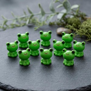 Puede incluir: Una colección de once pequeñas figuras de ranas de color verde brillante dispuestas en grupo. Cada rana presenta dos ojos negros y una línea roja fina y sencilla para la boca. Las figuras son lisas y tienen una forma redondeada y compacta.