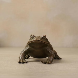 May include: A bronze-colored toad figurine with a textured, bumpy surface and a wide, flat mouth. The sculpture is depicted in a low, crouching position with its front legs extended forward, showcasing detailed skin patterns and defined eyes.