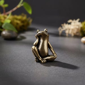 Op de afbeelding: Een klein koperen kikkerbeeldje in antieke stijl, afgebeeld in een zittende, contemplatieve houding met één hand rustend bij de kin. Het metalen oppervlak heeft een donkere, gebruinde patina met accenten op de ledematen en het hoofd.