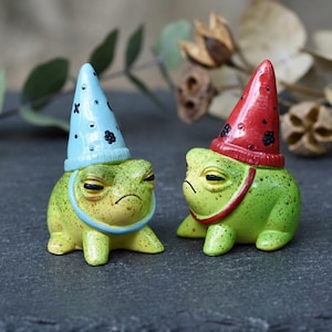 Puede incluir: Dos pequeñas figuras de ranas verdes con piel moteada y expresión gruñona. Una rana lleva un sombrero puntiagudo azul claro con símbolos negros y la otra un sombrero puntiagudo rojo con motivos florales negros, ambos sujetos con correas.