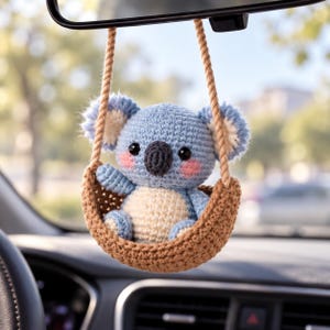 May include: A crocheted koala ornament hanging from a rearview mirror. The koala has light blue fur, a cream-colored belly, dark eyes, a dark gray nose, and pink cheeks. It sits inside a small, brown, bowl-shaped crocheted swing with rope.