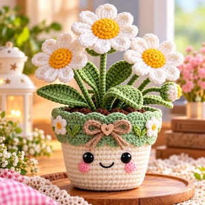 Puede incluir: Una planta en maceta tejida a ganchillo con tres margaritas blancas de centro amarillo y hojas verdes. La maceta color crema tiene un borde festoneado verde, un pequeño corazón de madera, un lazo de cuerda marrón y una cara bordada con ojos negros.
