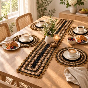 May include: A rectangular crochet table runner and four matching round crochet placemats in black, tan, and mustard yellow. The set is arranged on a wooden table with white ceramic coffee cups, saucers, and plates featuring croissants and berries.