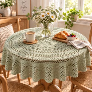 May include: A round, sage green crochet tablecloth with an intricate, openwork lace pattern featuring a scalloped edge. The tablecloth is draped over a circular wooden table, providing a textured, decorative covering for the tabletop surface.