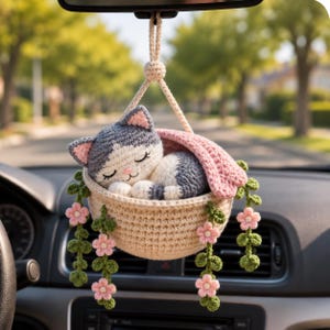 Könnte beinhalten: Ein gehäkeltes Auto-Hängeornament mit einer schlafenden grau-weißen Katze in einem beigen Korb. Der Korb ist mit grünen Ranken und kleinen rosa Blüten verziert und wird an einer beigen Kordel von einem zentralen Knoten über der Katze gehalten.