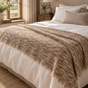 Peut inclure: Un jeté de lit rectangulaire présentant un tissage texturé aux nuances brunes et crème, agrémenté de courtes franges sur les bords, disposé au pied d'un lit recouvert de draps blancs et d'un coussin rectangulaire assorti de couleur brune.