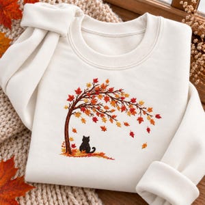 Pode incluir: Uma camisola de gola redonda cor de creme com um design bordado de um gato preto sentado debaixo de uma árvore curva com folhas de outono a cair em tons de laranja, vermelho e amarelo. A camisola tem mangas compridas e gola canelada.