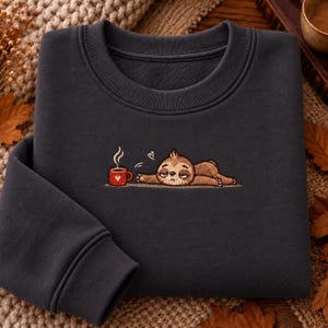 May include: A dark charcoal grey crewneck sweatshirt featuring an embroidered graphic of a brown sloth lying down next to a red mug with a white heart. The sloth appears tired with half-closed eyes, and a small white heart floats above its head.