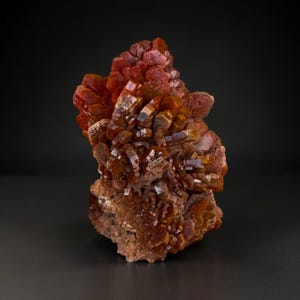 May include: A cluster of vibrant, deep orange-red vanadinite crystals featuring hexagonal, tabular structures. The mineral specimen displays a dense arrangement of sharp, glossy crystal faces emerging from a rough, earthy, brownish-red matrix base.