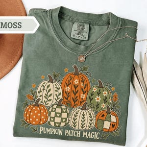 May include: A moss green t-shirt featuring a graphic of seven pumpkins with various patterns including polka dots, checkers, and floral designs. The text PUMPKIN PATCH MAGIC is printed in white below the pumpkins. The shirt has a crew neckline.