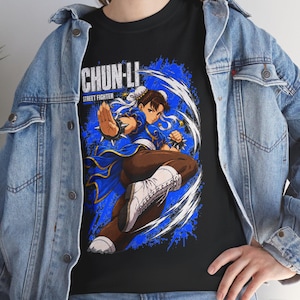 Puede incluir: Una camiseta negra con un estampado gráfico del personaje Chun-Li de Street Fighter en una pose dinámica de artes marciales, vistiendo un traje azul con botas blancas, bajo una chaqueta vaquera azul claro y pantalones vaqueros.
