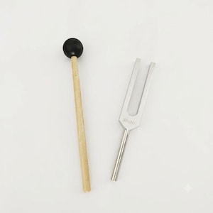 May include: A silver-coloured metal tuning fork engraved with the text 888Hz, positioned next to a wooden mallet featuring a smooth, round black rubber head. The tuning fork has a two-pronged design with a long, cylindrical handle for holding.