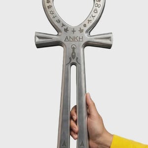 Könnte beinhalten: Ein metallisches, silberfarbenes Ankh-Symbol mit einem kreisförmigen Oberteil und eingravierten Symbolen am Rand. Das Wort ANKH ist in der Mitte aufgedruckt, ergänzt durch lineare Gravuren und geometrische Muster an den zwei vertikalen Beinen.