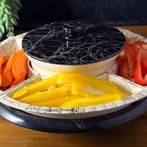 May include: A multi-compartment serving dish with a black and white patterned lid. The dish contains orange carrots, yellow peppers, and red bell peppers. The dish sits on a dark blue base.