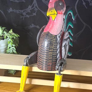 May include: A hand-painted wooden cockerel figurine with a red head featuring white polka dots, a yellow beak, and a black body with horizontal orange stripes. The cockerel has long, articulated yellow legs with webbed feet and black and green tail feathers.