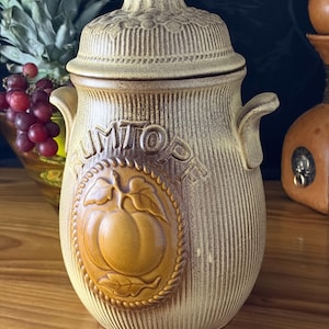 May include: A tan ceramic Rumtopf jar with a lid, featuring a vertical ribbed texture and a central embossed oval medallion depicting a piece of fruit with leaves. The jar has two side handles and a textured lid with a knob-style handle on top.
