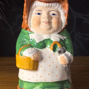 Peut inclure: Une cruche en céramique en forme de personnage portant un bonnet marron, une robe verte et un tablier blanc à pois orange. La figurine tient un panier jaune dans une main et un parapluie gris dans l'autre, avec des yeux bleus et un col blanc.