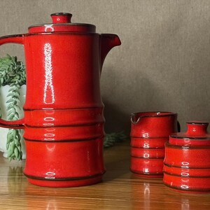 May include: A red ceramic coffee set comprising a coffee pot, creamer, and sugar bowl. The set features a textured surface and black trim. The coffee pot has a handle and lid. The creamer has a spout, and the sugar bowl has a lid.