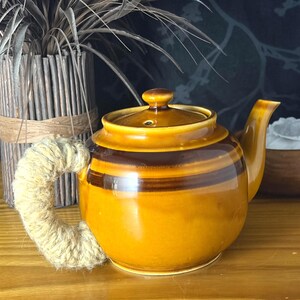 May include: A ceramic teapot featuring a glossy mustard yellow and dark brown horizontal striped glaze. The teapot has a rounded body, a matching lid with a small knob, and a curved handle wrapped in thick, textured, light beige natural fibre twine.