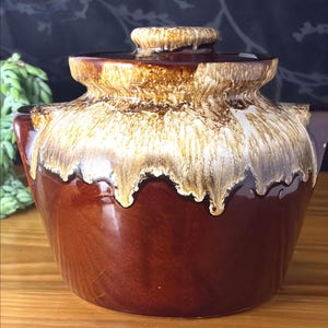 May include: A ceramic lidded jar featuring a deep brown base colour with a contrasting cream-coloured glaze that drips down from the rim. The lid has a matching brown and cream drip glaze finish with a central knob handle on the top surface.