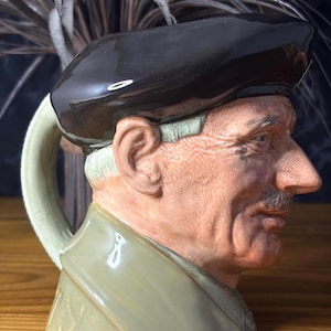May include: A ceramic character jug shaped as the head and shoulders of an older person wearing a dark brown beret and a light olive green collared coat. The figure has a textured skin tone, a mustache, and a handle attached to the back of the neck.