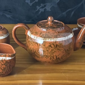 May include: A ceramic tea set featuring a teapot, a lidded sugar bowl, a creamer, and a teacup with a saucer. Each piece has a glossy brown glaze with dark brown mottled patterns and a distinct horizontal white band encircling the middle section.