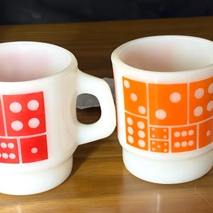 May include: Two white milk glass mugs featuring a stacked design. One mug displays a red domino pattern, while the other features an orange domino pattern. Both mugs have a cylindrical body with a thick handle and a slightly recessed base.