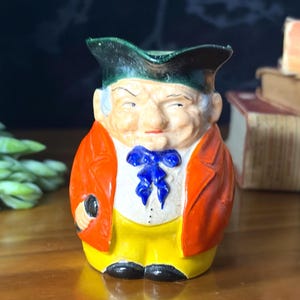 Peut inclure: Une cruche en céramique en forme de personnage portant un tricorne vert, une veste orange, un gilet blanc avec une cravate bleue et un pantalon jaune. La figurine présente des traits peints et tient un petit objet noir dans une main.