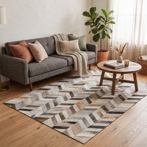May include: A rectangular area rug featuring a multi-colored chevron pattern made of stitched hide panels in shades of gray, brown, white, and tan. The rug is positioned on a light wood floor in front of a gray sofa with various throw pillows.