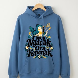 Peut inclure: Un sweat à capuche bleu poussiéreux orné d'un motif représentant un oiseau bleu chanteur avec des accents jaunes. Sous l'oiseau, le texte Ora Ngapak Ora Kepenak est écrit en caractères sombres, entouré de fleurs blanches et de feuilles vertes.