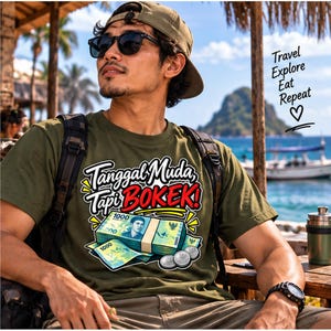 May include: An olive green t-shirt featuring a graphic print of a stack of 1000 Indonesian Rupiah banknotes and coins. The text reads "Tanggal Muda, Tapi BOKEK!" in white and red lettering, with "Travel Explore Eat Repeat" and a heart symbol.