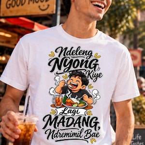 Op de afbeelding: Een wit T-shirt met korte mouwen en een grafische illustratie van een persoon die een maaltijd met rijst, kip en een drankje eet. De tekst luidt Ndeleng Nyong Kiye Lagi MADANG, Ndremimil Bae in zwarte en gele gestileerde letters.