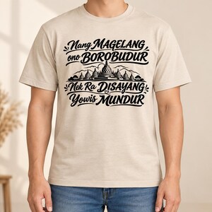 May include: A beige short-sleeved t-shirt featuring black graphic text that reads Nang MAGELANG ono BOROBUDUR Nek Ra DISAYANG Yowis MUNDUR, centered above a detailed black illustration of the Borobudur temple complex with surrounding trees and mountains.