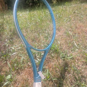 Op de afbeelding: Een blauwgroen Head tennisracketframe met een witte grip en een witte rand langs de binnenkant van het blad. Het frame toont de merknaam Head in kleine witte letters bij de keel, met een klein wit label bevestigd aan de schacht.