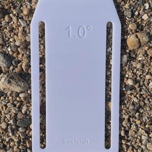 Puede incluir: Una herramienta rectangular de plástico blanco con esquinas superiores redondeadas y dos ranuras verticales largas. El texto 1.0° está grabado cerca de la parte superior y la palabra Swellcraft está grabada cerca de la base. Superficie lisa y plana.