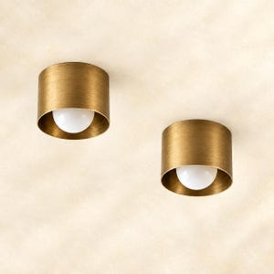 May include: Two cylindrical flush mount ceiling lights featuring a brushed brass finish on the exterior and interior. Each fixture contains a single, centered, spherical white glass bulb that emits light from within the metal housing.