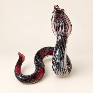 May include: A glass snake figurine featuring a coiled body and an upright head. The glass is deep red and black with white swirling patterns and fine glittery flecks throughout the surface, creating a textured appearance across the entire sculpture.