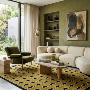 Op de afbeelding: Een woonkamer met een crèmekleurige, gebogen bank met twee ronde kussens, een donker olijfgroene fluwelen fauteuil en een rechthoekige salontafel met een blad van natuursteen. Een geel en zwart vloerkleed met geometrisch patroon.