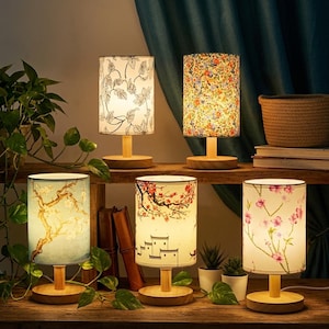 May include: Five cylindrical table lamps with light wood bases and fabric shades. The shades feature various prints including black and white leaves, colorful floral patterns, a blue tree branch, a landscape with buildings, and pink cherry blossoms.