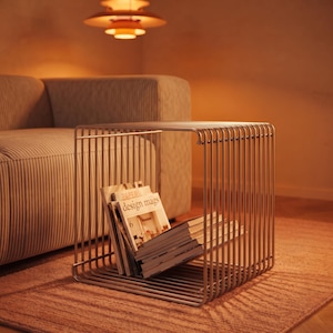 Può includere: Un tavolino a forma di cubo costruito con sottili aste metalliche parallele in una calda finitura metallica. Il design a telaio aperto presenta una superficie superiore piatta e una base che contiene diverse riviste, tra cui una con il testo design mags.
