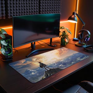May include: A rectangular desk mat featuring a printed landscape illustration of a figure standing on a rocky cliff overlooking a vast mountain range at sunset. The mat has a smooth surface and is positioned on a dark desk surface in front of a monitor.