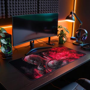 May include: A rectangular desk mat featuring a graphic print of the character Deadpool in a red and black color scheme with smoky effects. The mat is positioned on a dark desk surface in front of a curved computer monitor, a speaker, and headphones.