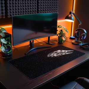 Op de afbeelding: Een rechthoekige zwarte bureaumat met een wit sterrenveldpatroon en een gedetailleerde afbeelding van een ruimteschip in het midden. De mat ligt op een donkerhouten bureau voor een gebogen computermonitor, een luidspreker en een koptelefoon.