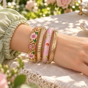 May include: A set of four gold-tone metal bangles worn on the wrist. One features an intricate openwork floral design with pink and green enamel, one is a plain textured band, one is a smooth pink enamel band with floral motifs, and one is engraved.